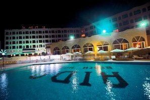 Un hotel con piscina, mobiliario exterior y un edificio grande de varios pisos y ventanales arqueados.