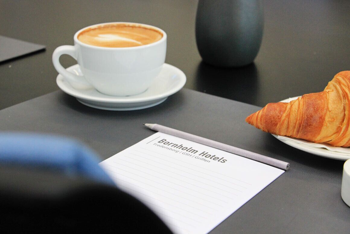 A table with a cup of coffee, a croissant, a notepad with "Bornholm Hotels" text, and a pen.