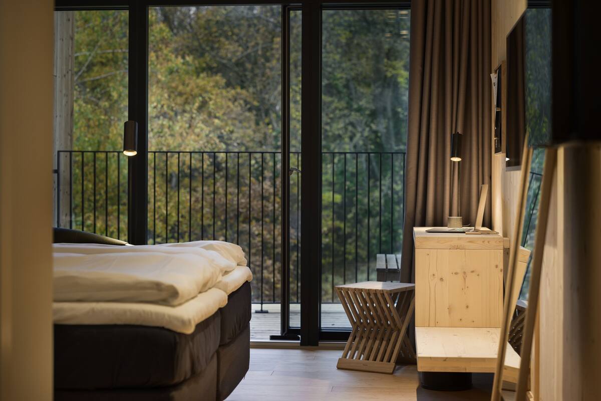 A bedroom with a large window, a bed, a small table, and a wooden cabinet.