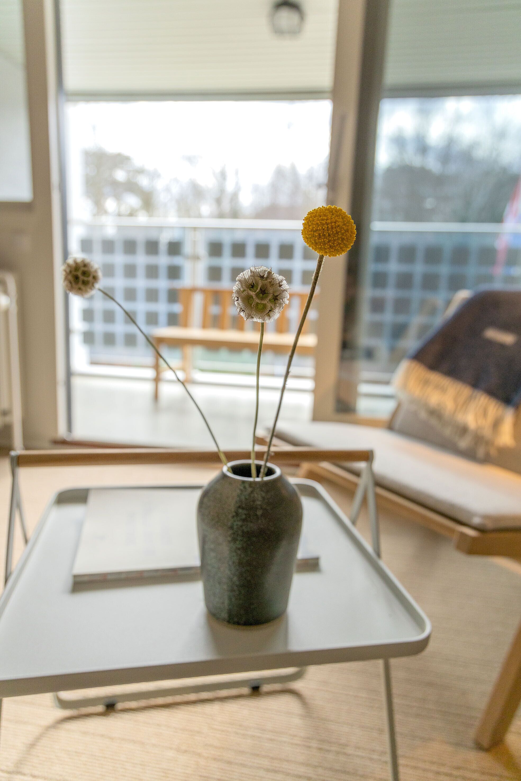 A table with a vase containing flowers, a chair, and a view of a balcony with a railing.