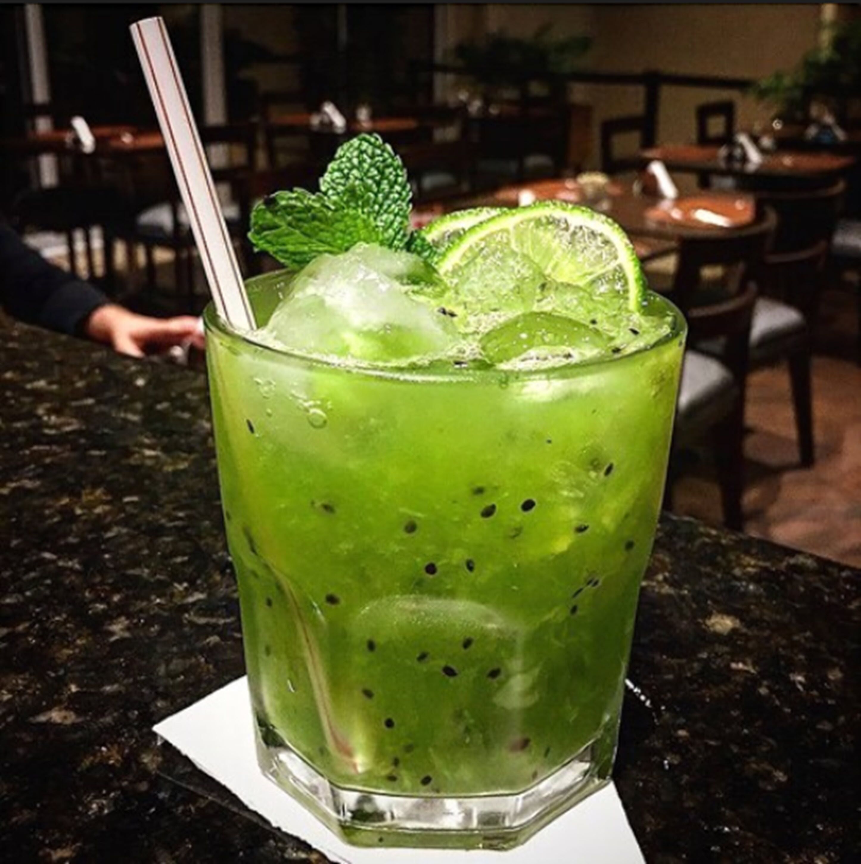 A glass of green beverage with ice and lime slices, garnished with mint, on a table.