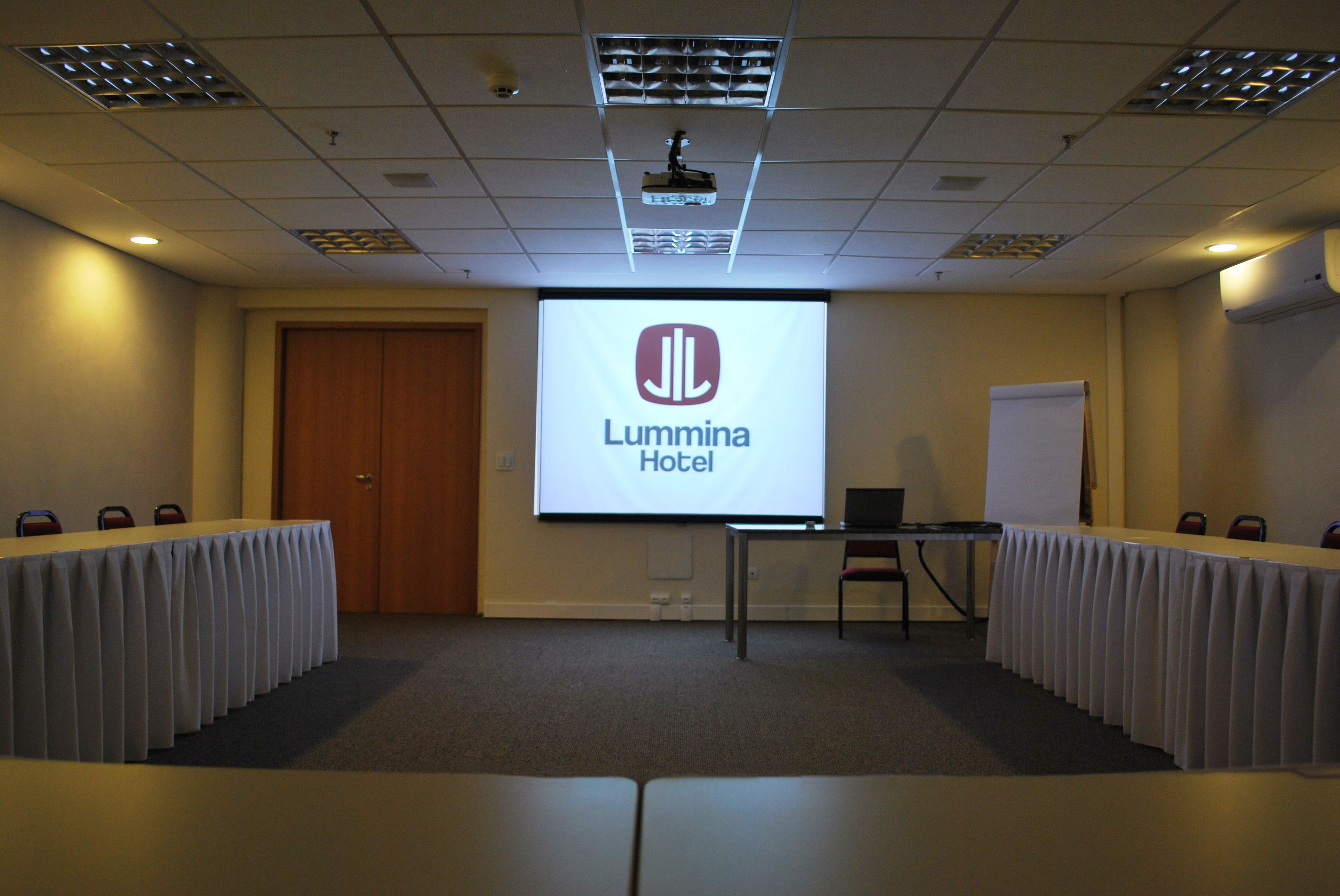Uma sala de conferências com tela de projeção exibindo o logotipo do Lumina Hotel, um quadro branco, um laptop e uma mesa com cadeiras.