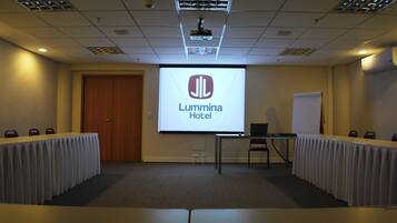 Uma sala de conferências com tela de projeção exibindo o logotipo do Lumina Hotel, um quadro branco, um laptop e uma mesa com cadeiras.