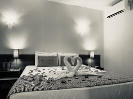 A hotel room with a bed featuring a patterned bedspread, two decorative pillows, a heart-shaped towel arrangement, and bedside lamps.