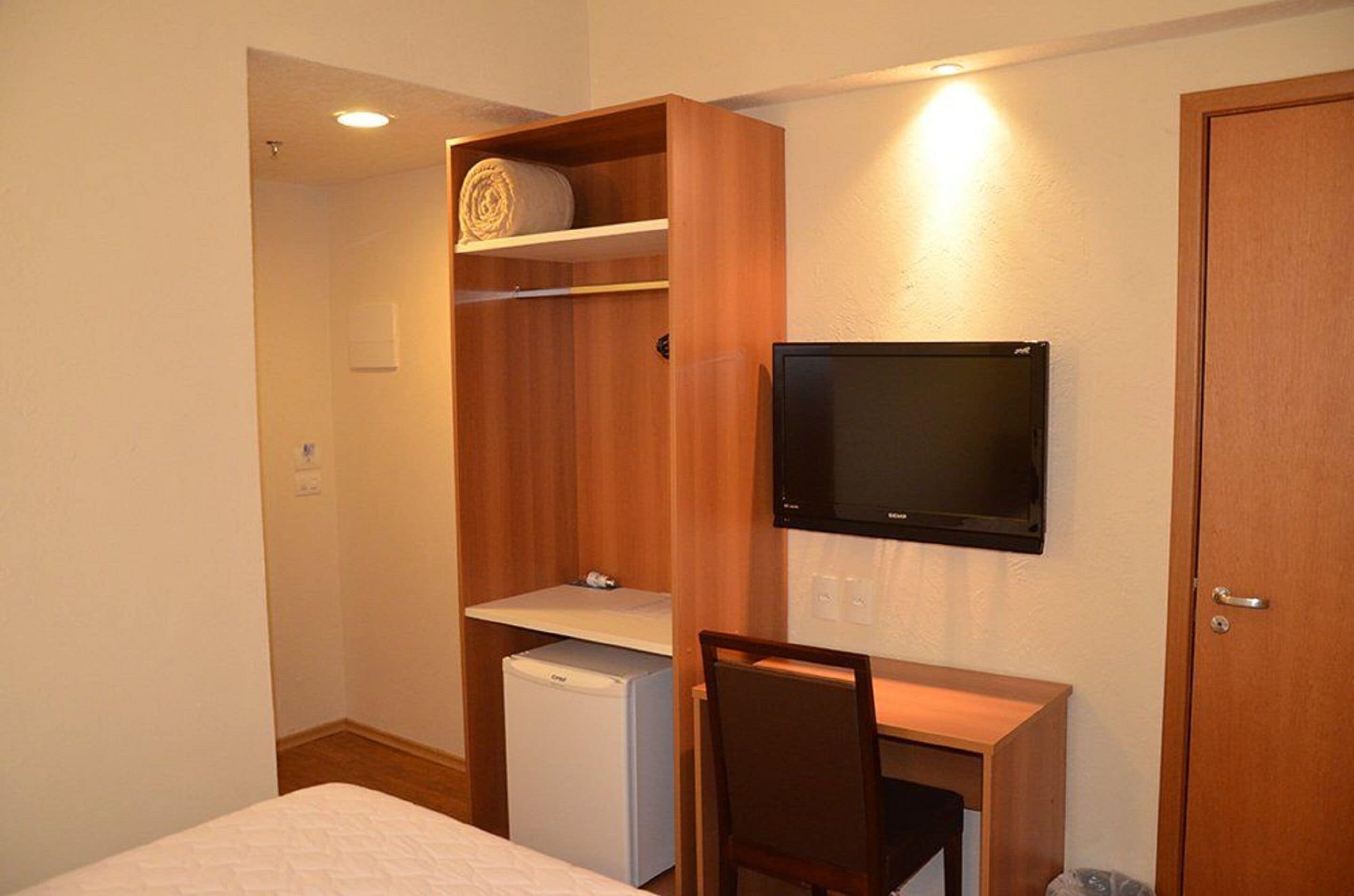 A compact room with a bed, a small desk with a chair, a television mounted on the wall, a wooden wardrobe, and a compact refrigerator.
