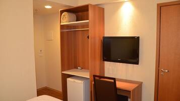 A compact room with a bed, a small desk with a chair, a television mounted on the wall, a wooden wardrobe, and a compact refrigerator.