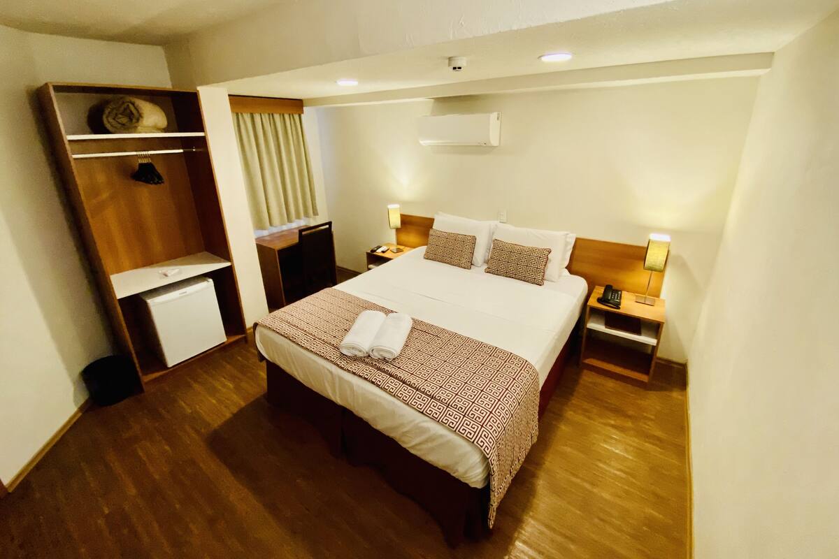 A hotel room with a large bed, a desk, a chair, a wardrobe, and a bedside table with a lamp.