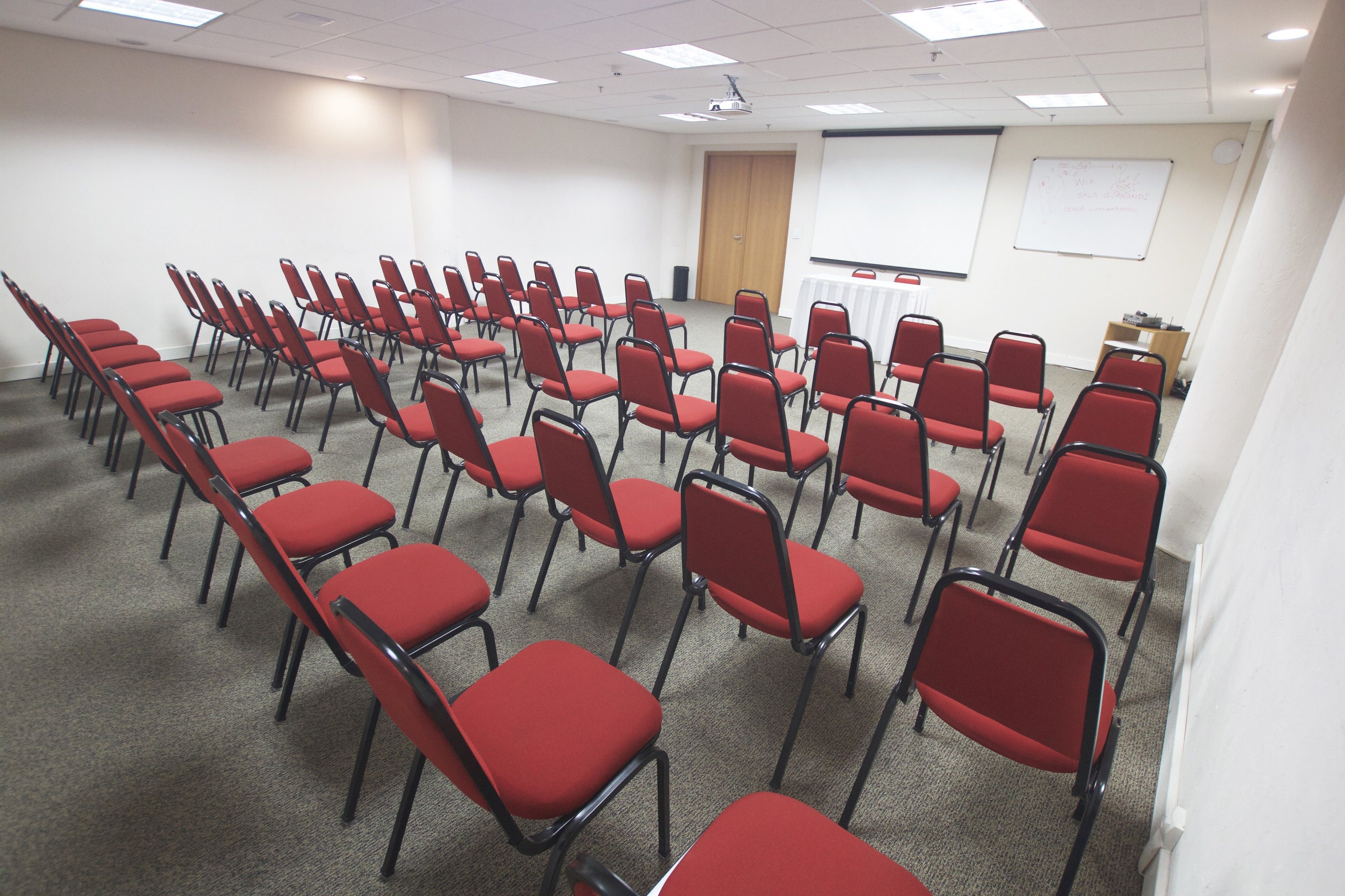 A conference room with rows of red chairs, a whiteboard, and a projector screen.