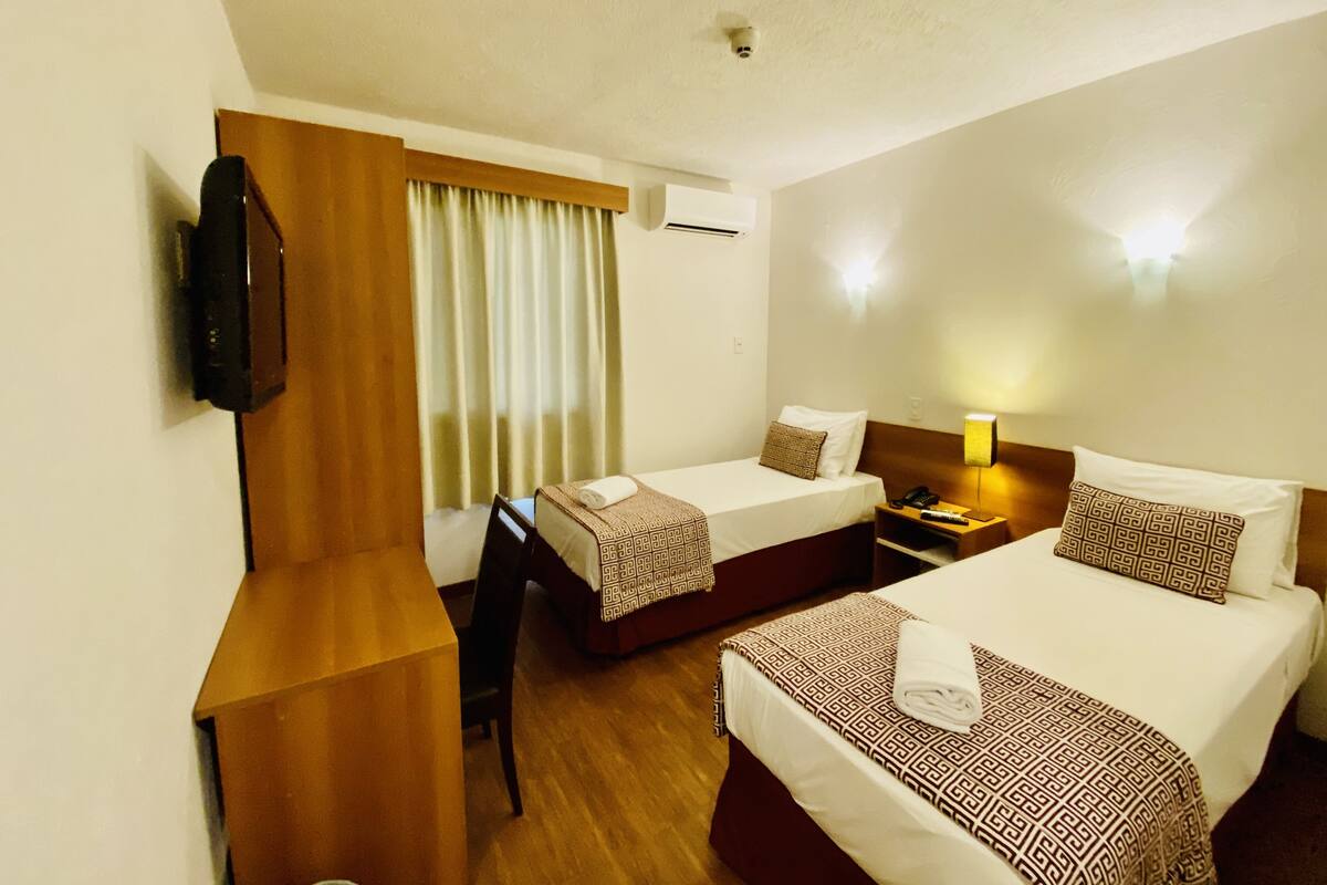A hotel room with two beds, a desk, a TV, and a window with curtains.