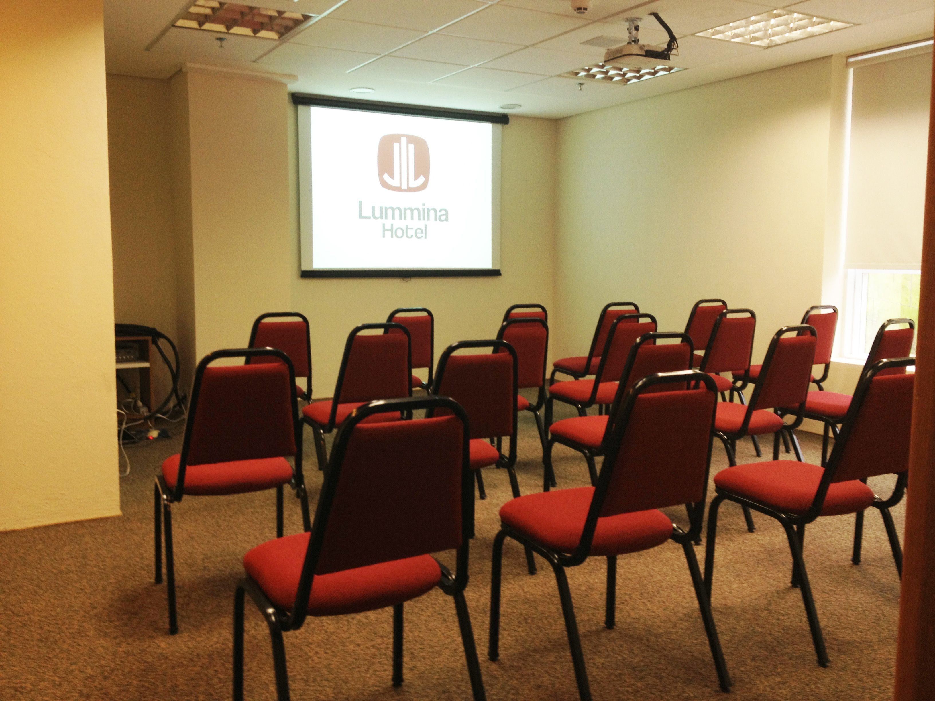 A conference room with red chairs arranged in rows, a projector screen displaying 'Lumma Hotel', and a ceiling with recessed lighting.