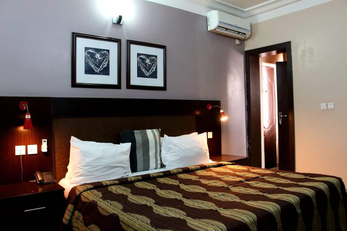 A hotel room with a bed, two framed pictures on the wall, a bedside lamp, a phone on the nightstand, and an air conditioning unit.