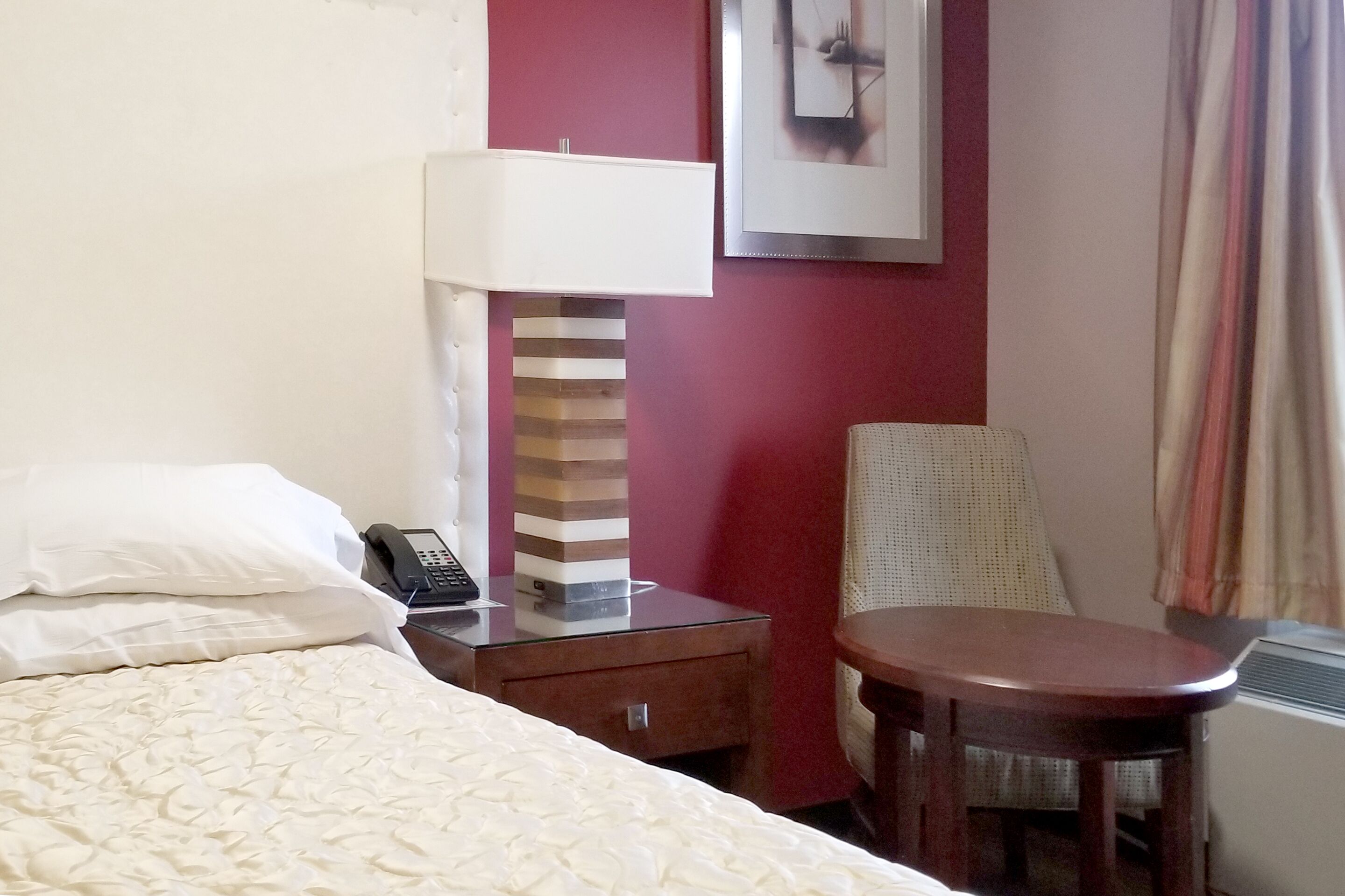 A hotel room with a bed, a bedside table with a lamp, a chair, and a framed picture on the wall.