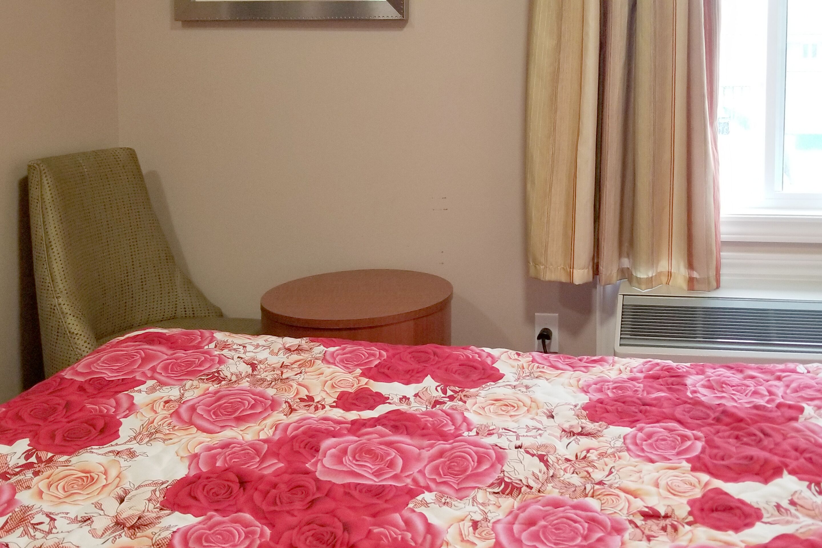 A hotel room with a floral bedspread, a round table, a chair, a window with curtains, and a wall-mounted air conditioning unit.