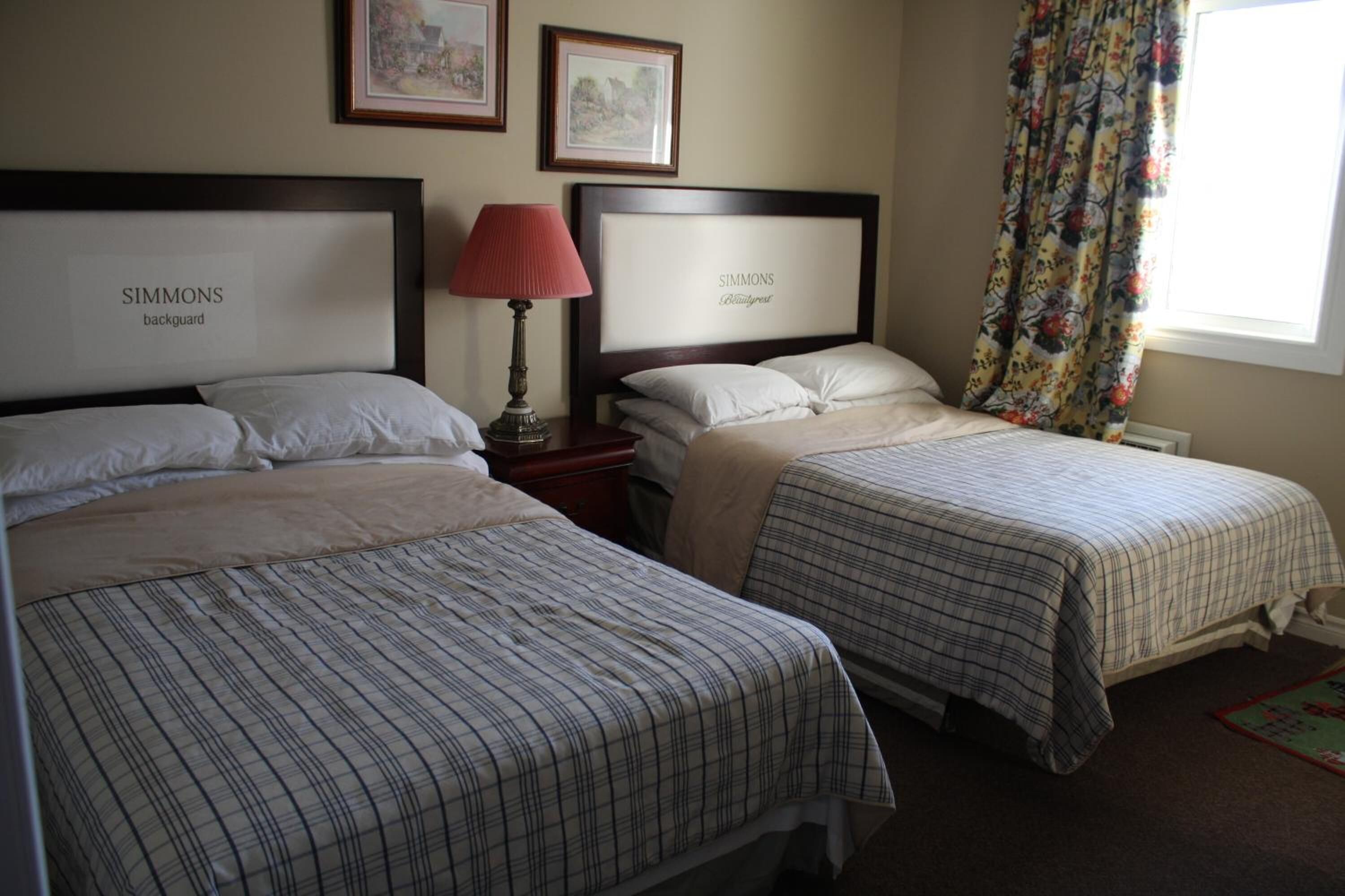A hotel room with two beds, each with a headboard labeled 'SIMMONS backguard'. There is a nightstand with a lamp, a window with floral curtains, and framed pictures on the wall.