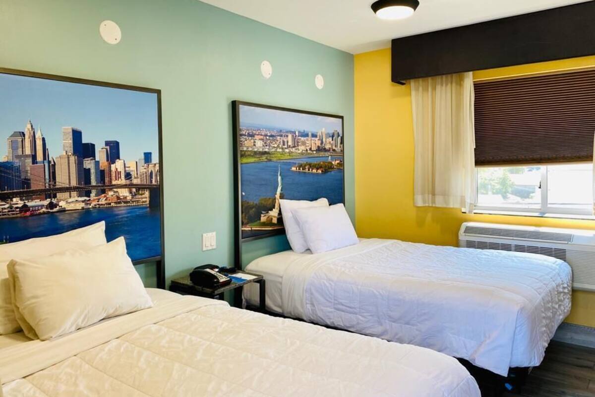 A hotel room with two beds, a cityscape mural on the wall, a window with blinds, and a ceiling light.