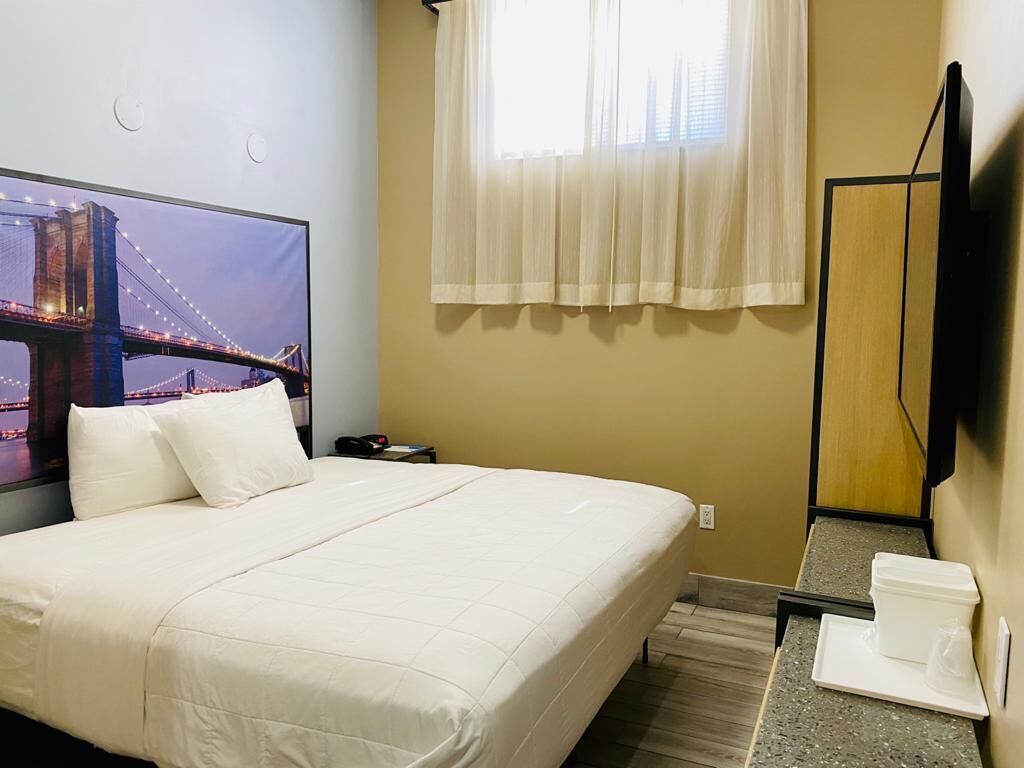 Habitación de hotel con cama, televisor mostrando una vista urbana, ventana con cortinas translúcidas y una pequeña mesa con bandeja.