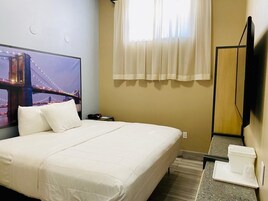 Habitación de hotel con cama, televisor mostrando una vista urbana, ventana con cortinas translúcidas y una pequeña mesa con bandeja.