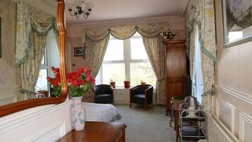 A room with a wooden dresser, a mirror, a vase with flowers, two chairs, a table with a tray, and a window with curtains.