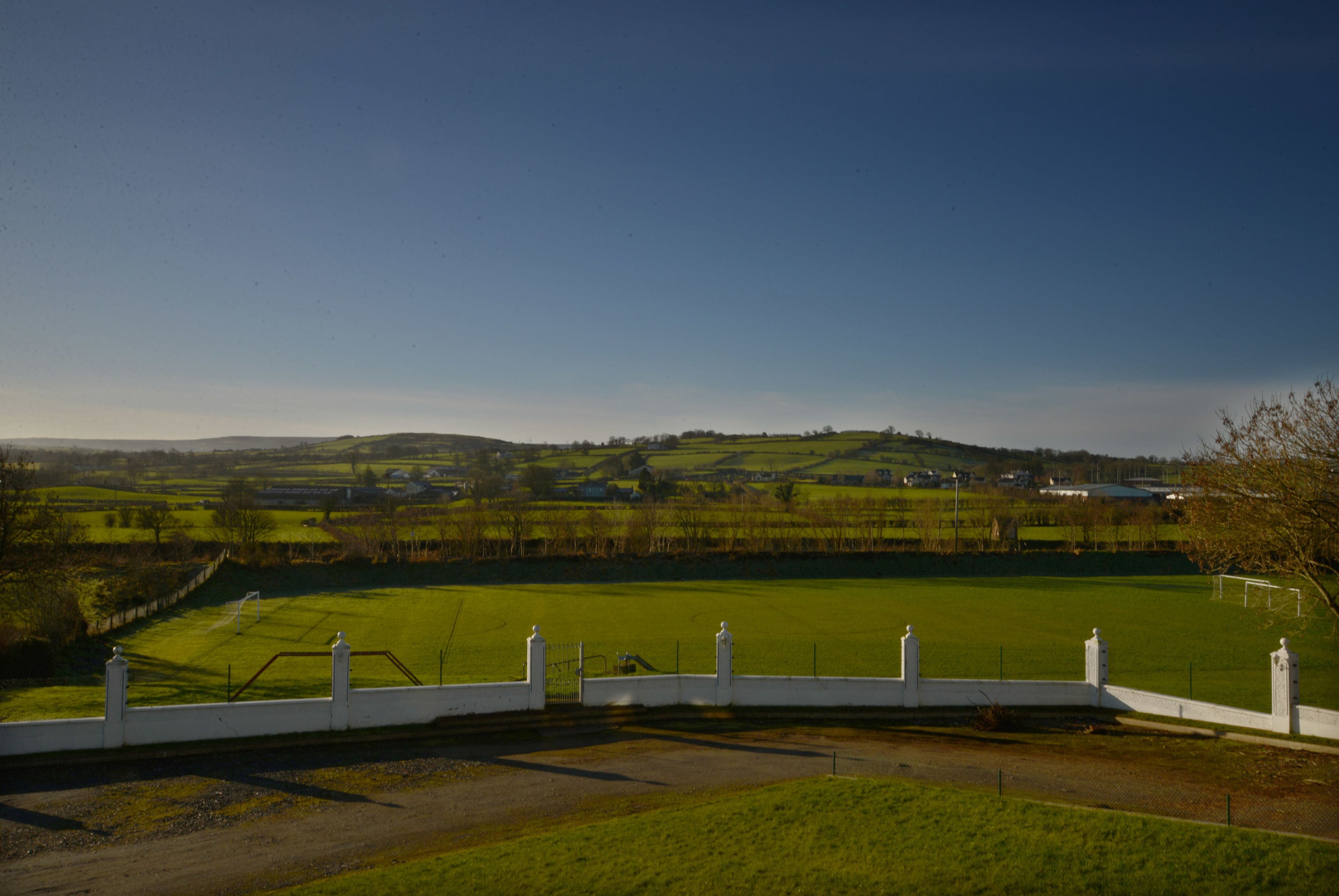 A rural landscape with a fenced field, a road, and distant buildings.