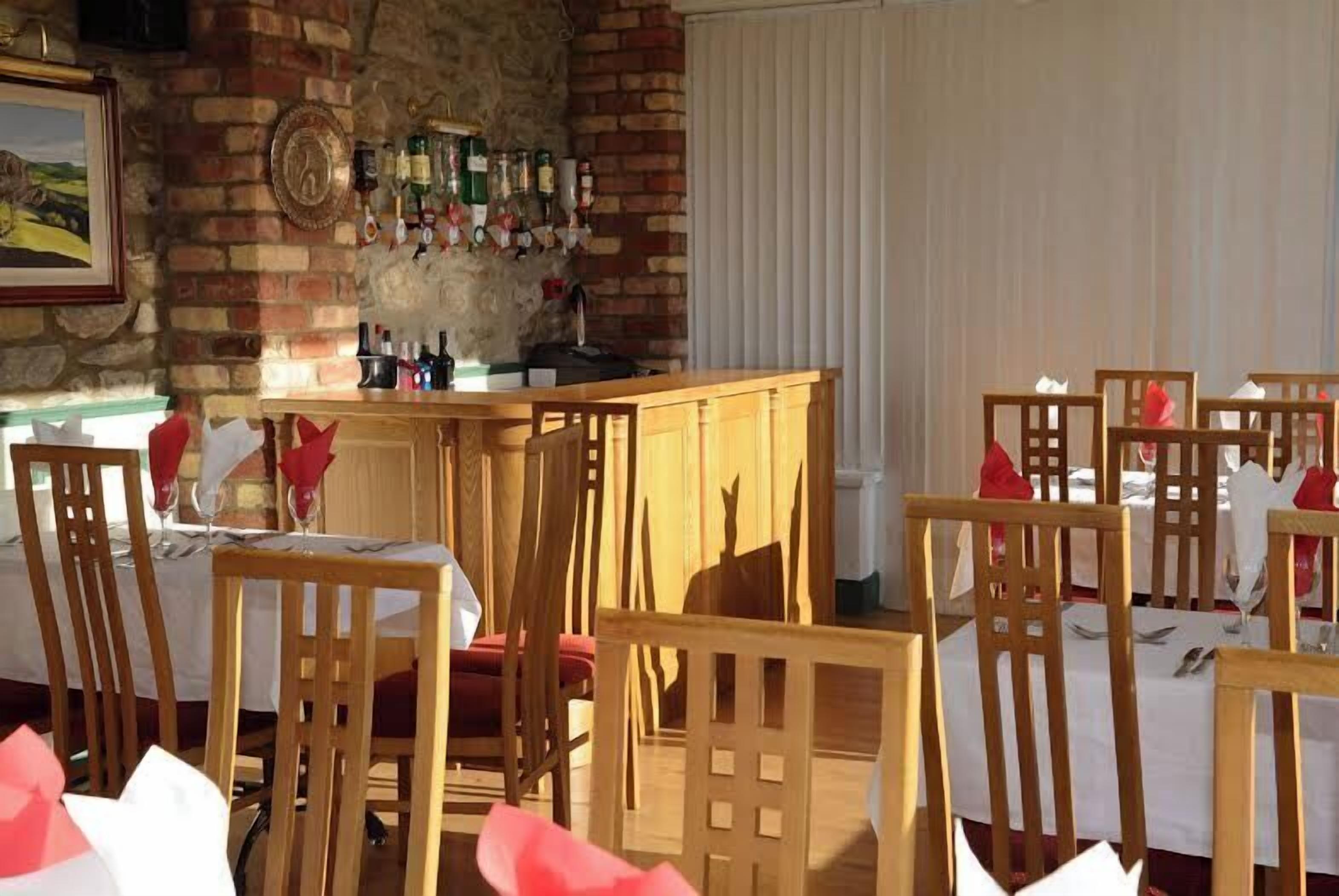 A dining area with wooden chairs and tables, a brick wall with a wine rack, and a window with blinds.