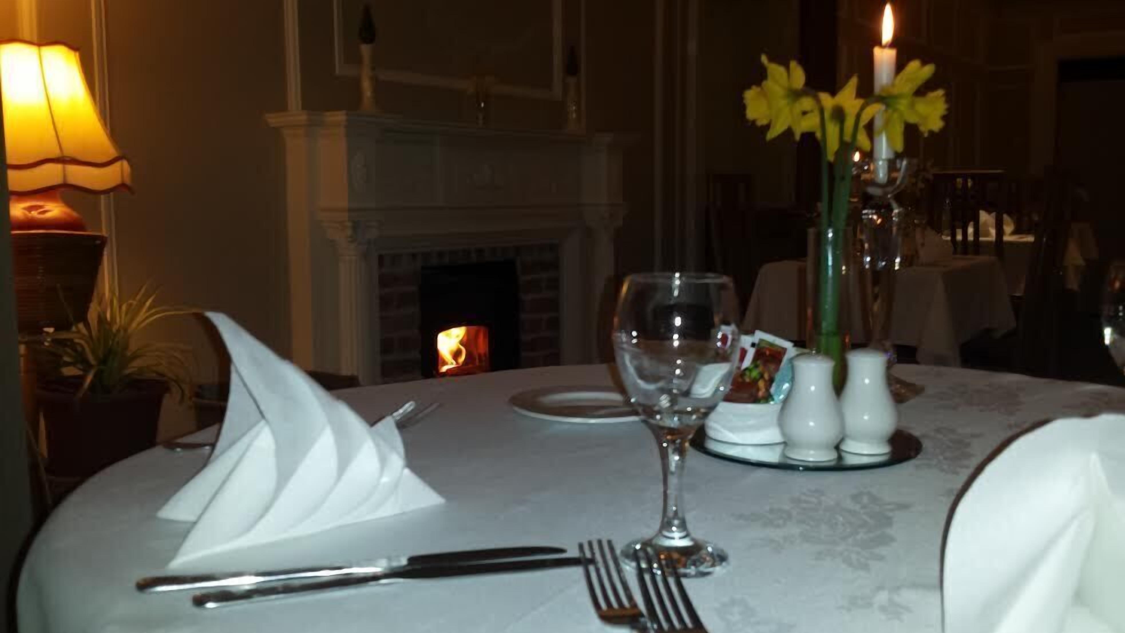 A dining table set for two with a white tablecloth, a vase of yellow flowers, salt and pepper shakers, and a lit fireplace in the background.