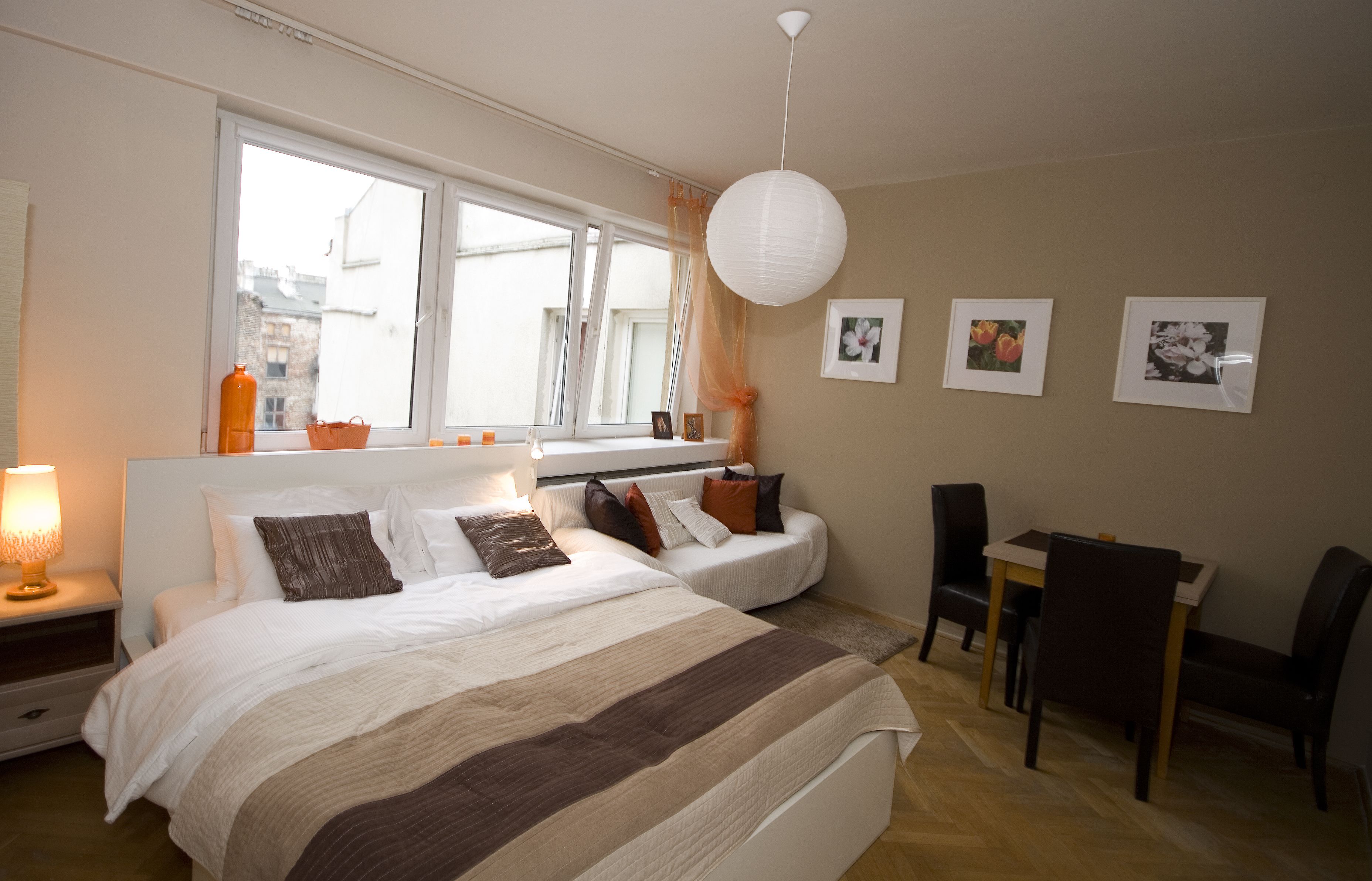 A bedroom with a large bed, bedside tables, a desk, and chairs. There are windows with curtains, a hanging light fixture, and framed pictures on the wall.