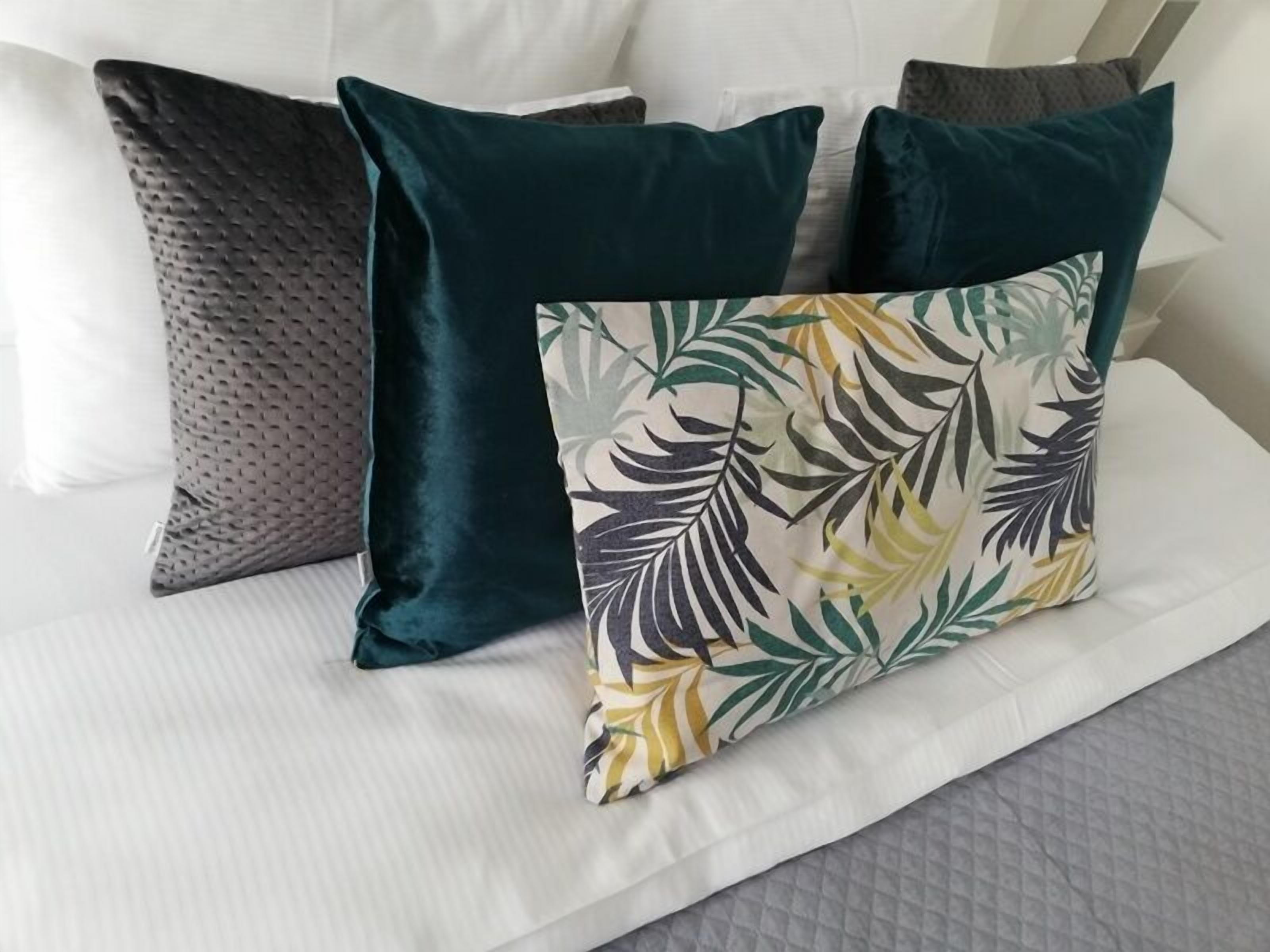 A bed with white bedding, a pillow with a tropical leaf pattern, and two dark teal pillows.