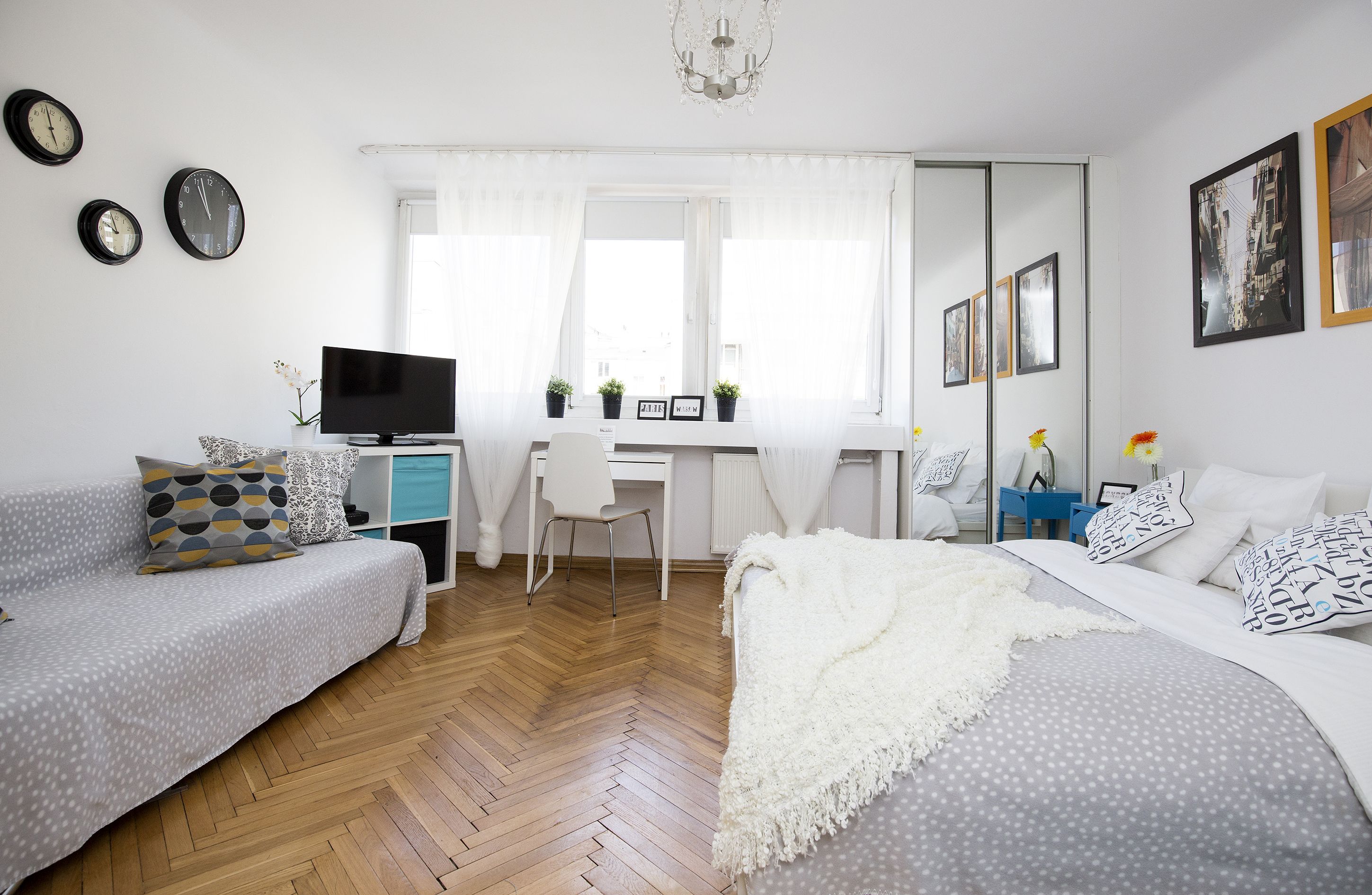 A modern bedroom with a wooden herringbone floor, a grey sofa with patterned cushions, a white bed with a fluffy white throw, a desk with a computer and two potted plants, and a large mirror.