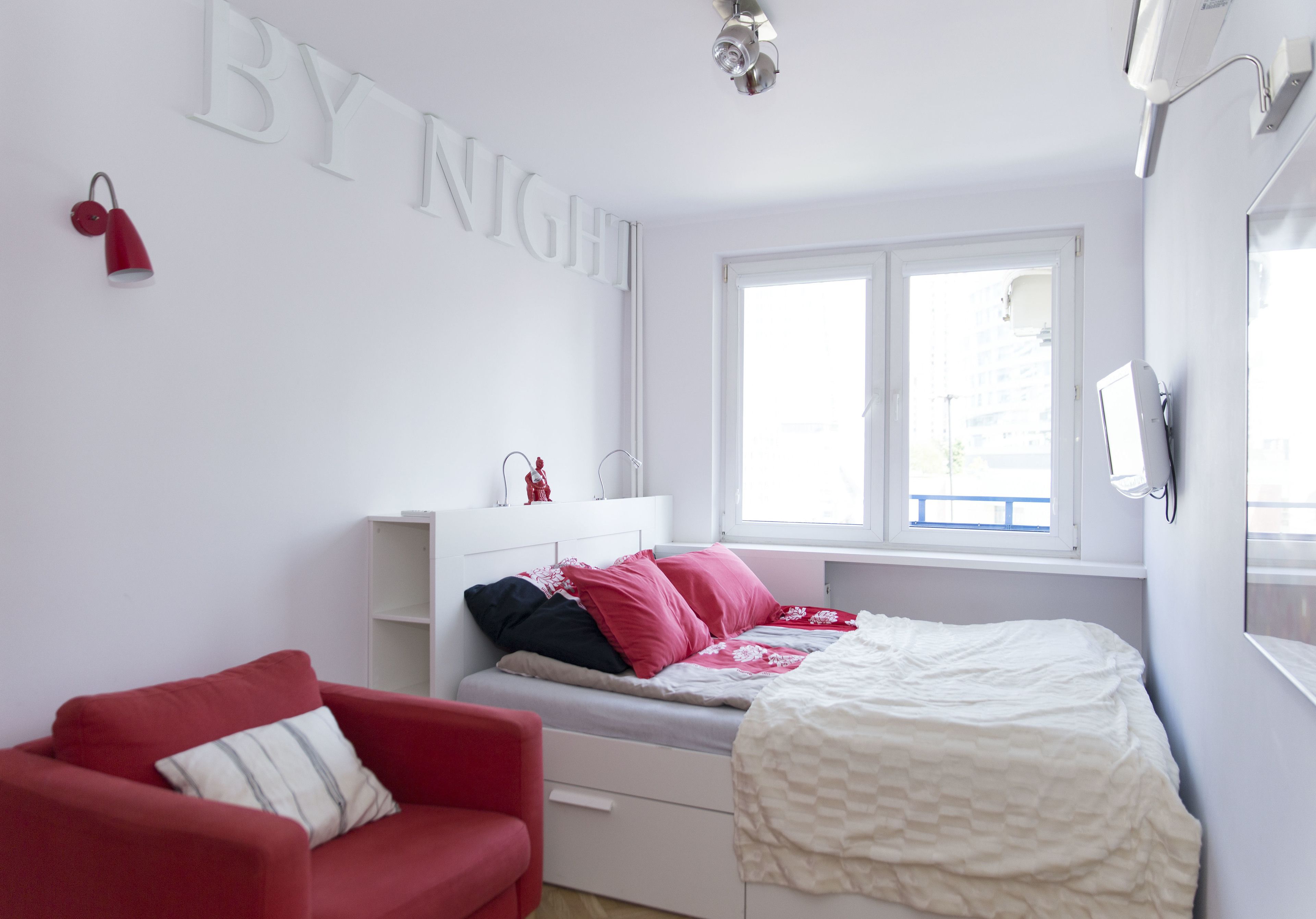 Une chambre à coucher avec un lit, un canapé rouge, une commode blanche et une fenêtre avec des stores.