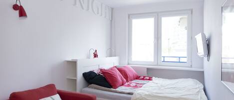 Une chambre à coucher avec un lit, un canapé rouge, une commode blanche et une fenêtre avec des stores.