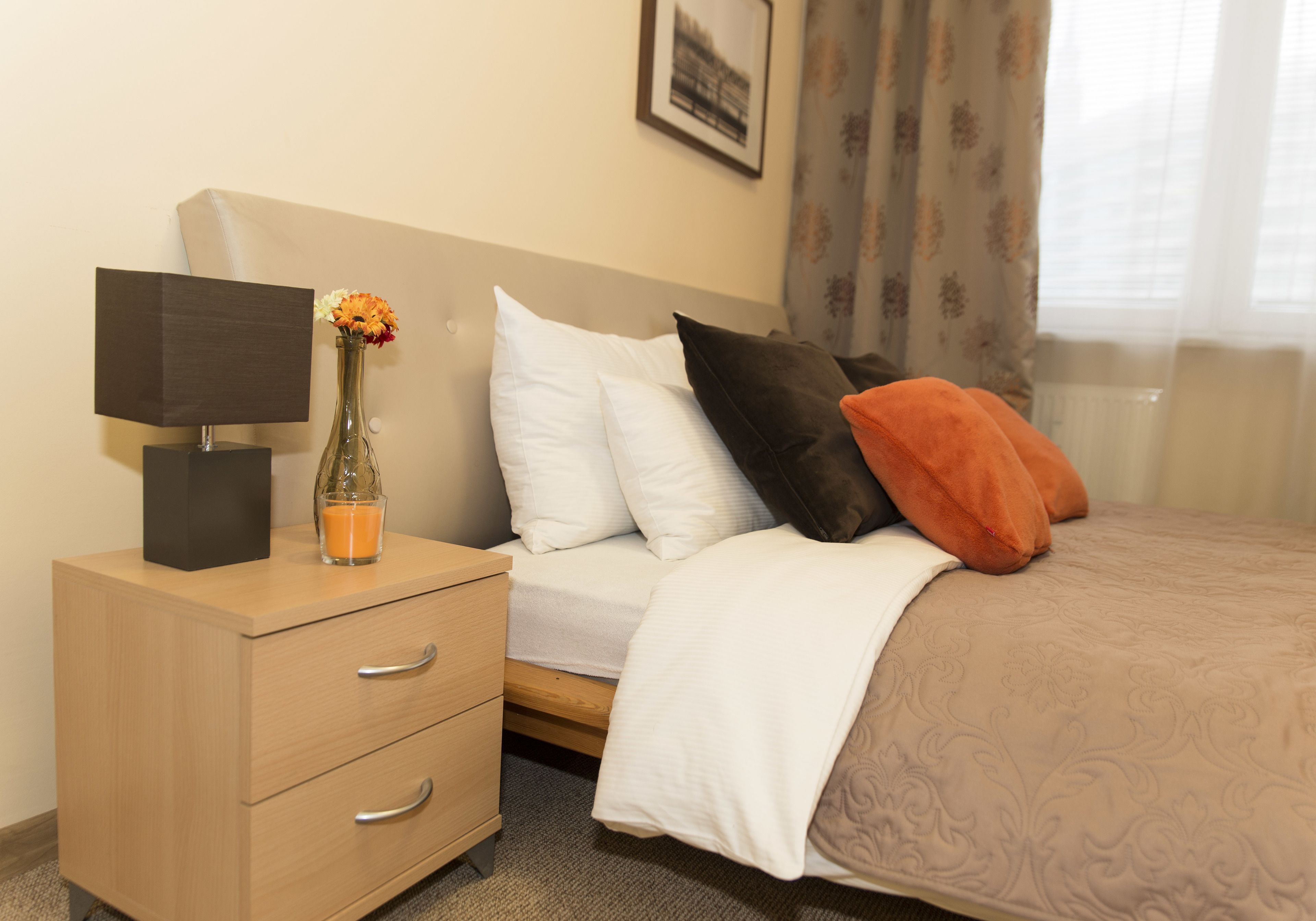 A neatly made bed with white and orange pillows, a bedside lamp, and a vase with flowers on a wooden nightstand.