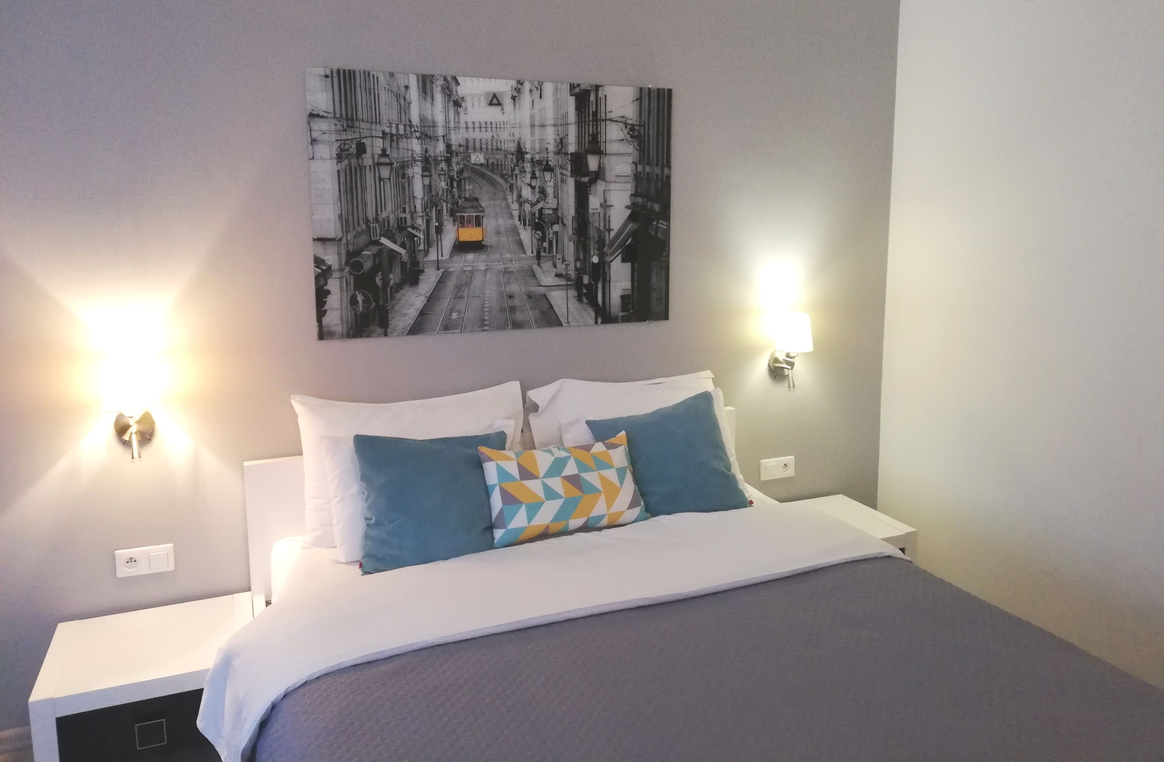 Una cama bien hecha con ropa de cama blanca y azul, un cojín de estampado geométrico y un cuadro de un paisaje urbano blanco y negro encima.