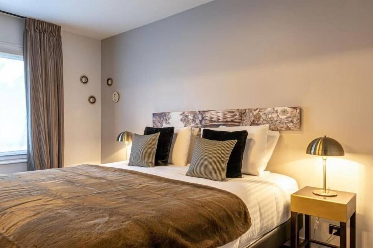 A hotel room with a large bed, a nightstand with a lamp, a wall clock, and a window with curtains.