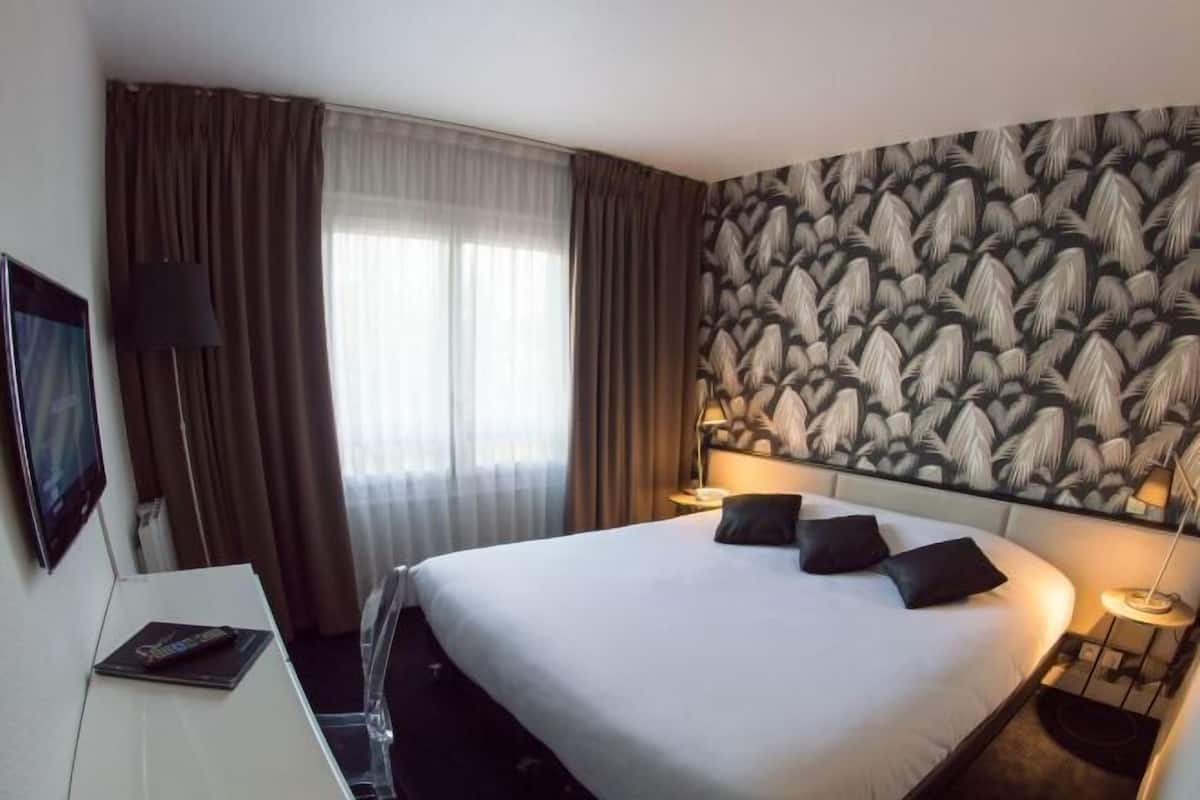 A hotel room with a bed, curtains, a headboard with a leaf pattern, a desk with a lamp, and a TV.