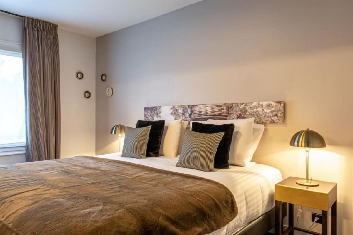 A hotel room with a large bed, a nightstand with a lamp, a wall clock, and a window with curtains.