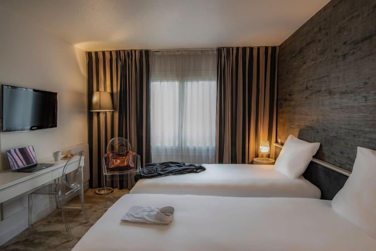 A hotel room with two beds, a desk with a laptop, a TV, and a window with curtains.