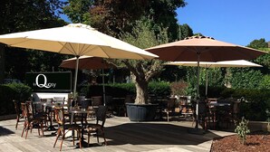 Un patio al aire libre con suelo de madera, sillas de mimbre y grandes sombrillas. Se ve un cartel con el nombre 'le Quosy'.