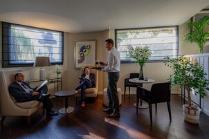 A modern interior with a seating area, a standing man holding a tray, and a large window with a view of greenery.