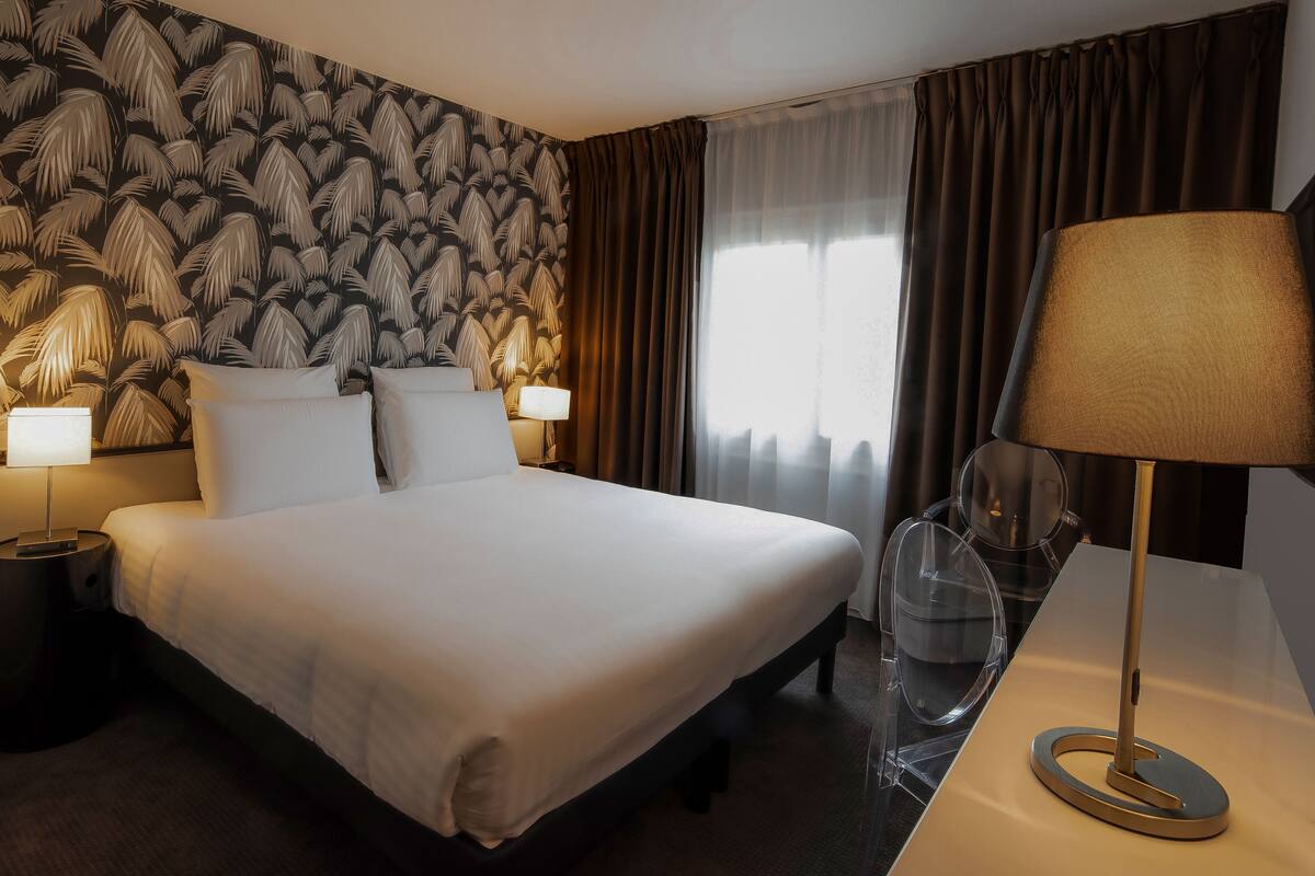 A hotel room with a large bed, two bedside tables with lamps, a desk with a lamp, and a window with curtains.
