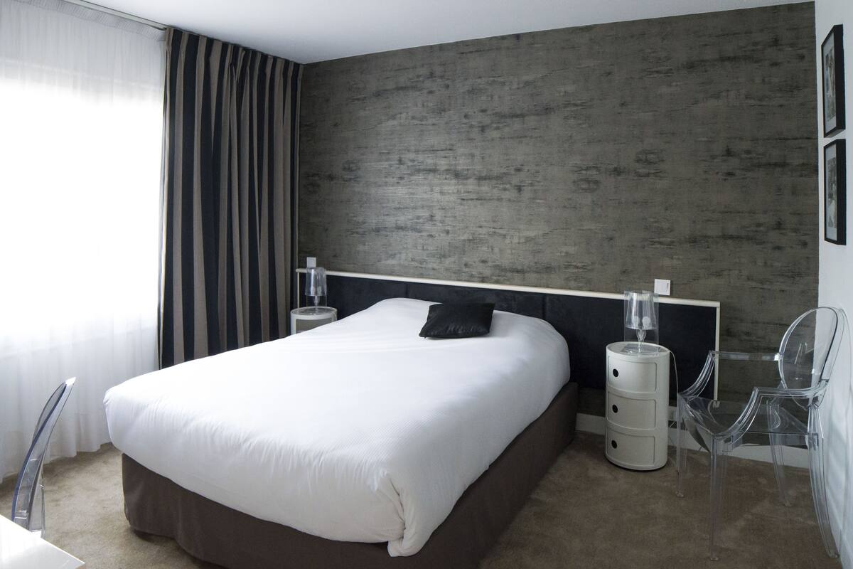 A modern hotel room with a large bed, a bedside table with a lamp, a clear chair, and a view of the city through the window.