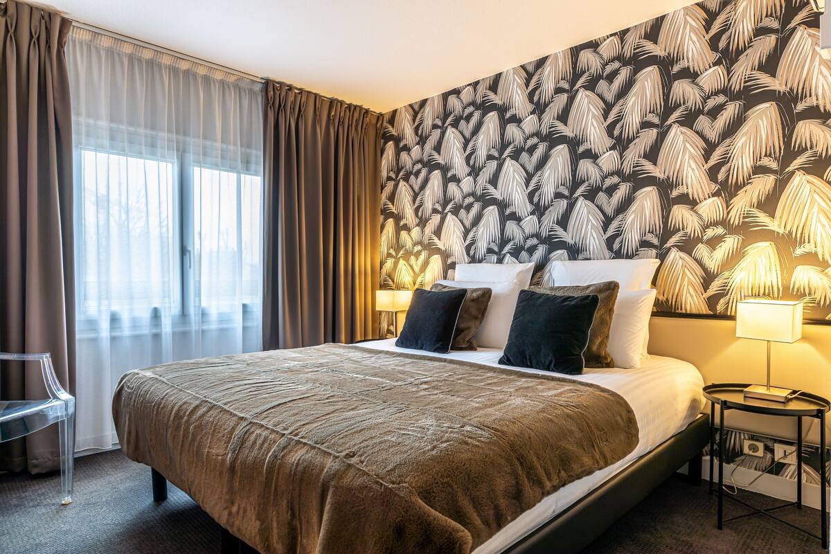 A hotel room with a large bed, a nightstand with a lamp, a chair, and a window with curtains.