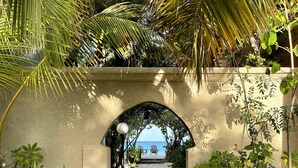 A concrete pathway leading through an arched structure surrounded by lush greenery and palm trees, with a view of the ocean in the distance. - Osprey Beach Hotel (Cockburn Town)