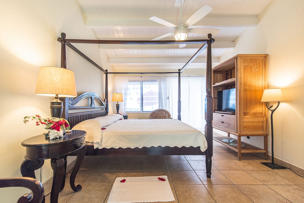 A four-poster bed with a canopy, a television on a wooden stand, a small table with a lamp, and a decorative vase with flowers.