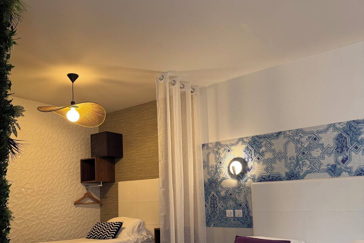 A hotel room with a large bed, a smaller bed, a ceiling fan, a wall-mounted light, a patterned wall, and a plant.