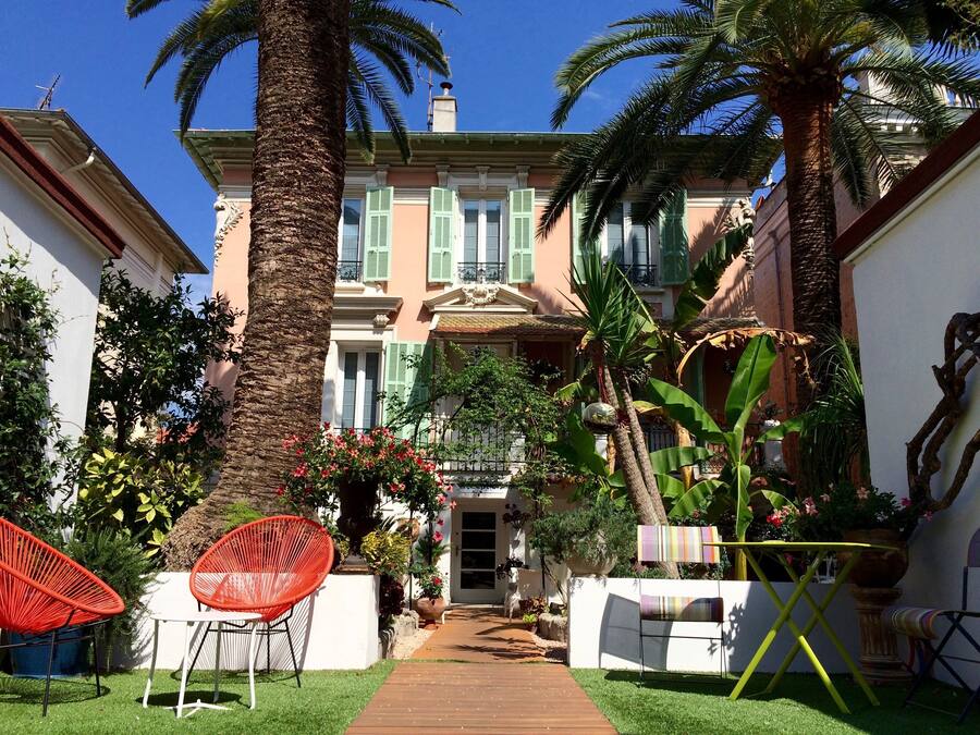Un patio con un sendero que lleva a un edificio con contraventanas verdes, rodeado de palmeras y plantas en macetas.