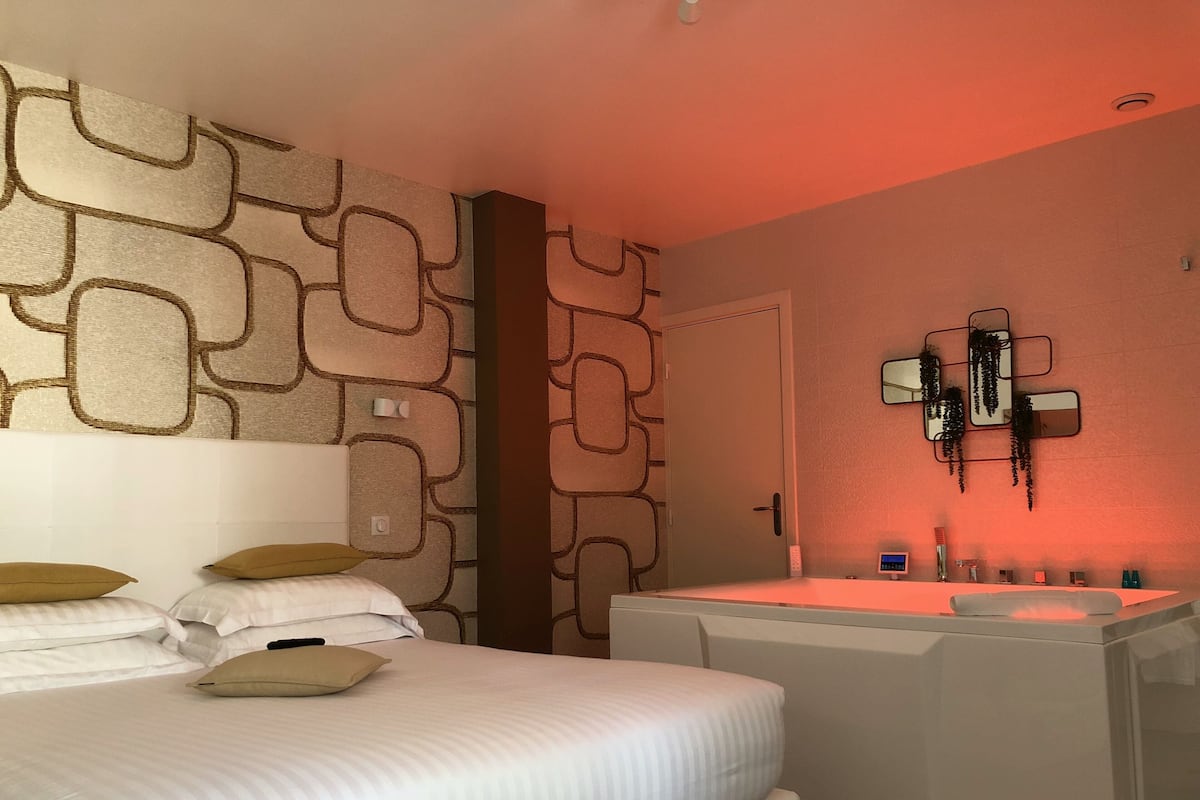 A hotel room with a bed, a bedside table, a mirror, a wall-mounted light, and a bathroom with a sink and mirror.