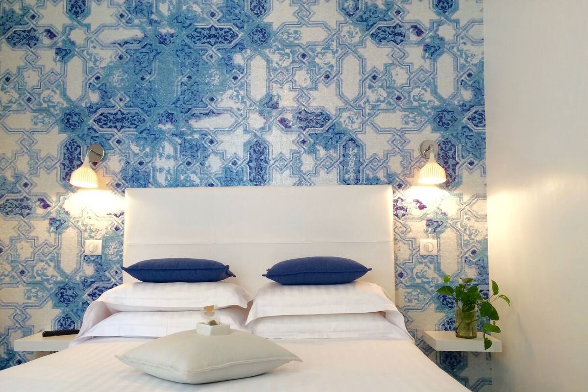 A neatly made bed with white linens and two blue pillows, a white headboard, and a wall with a blue geometric pattern.