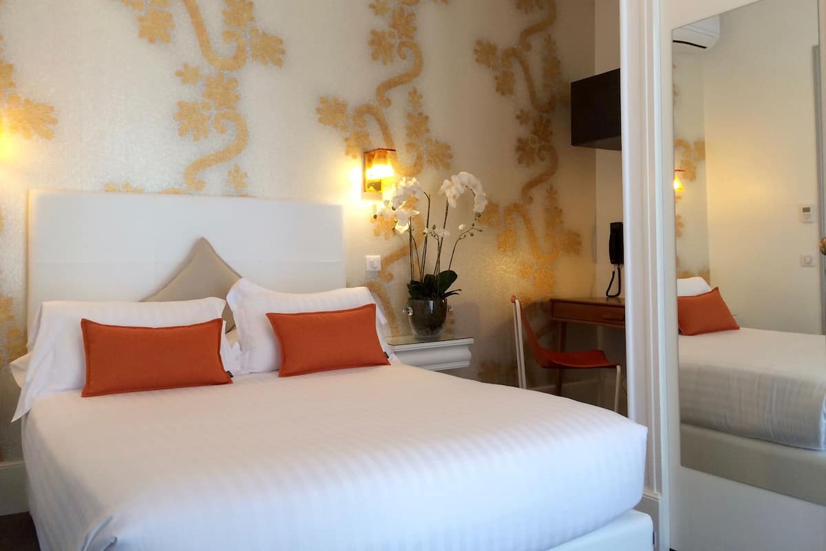 A hotel room with a large bed, white bedding, orange pillows, a bedside table with a lamp, a vase with flowers, and a mirror.