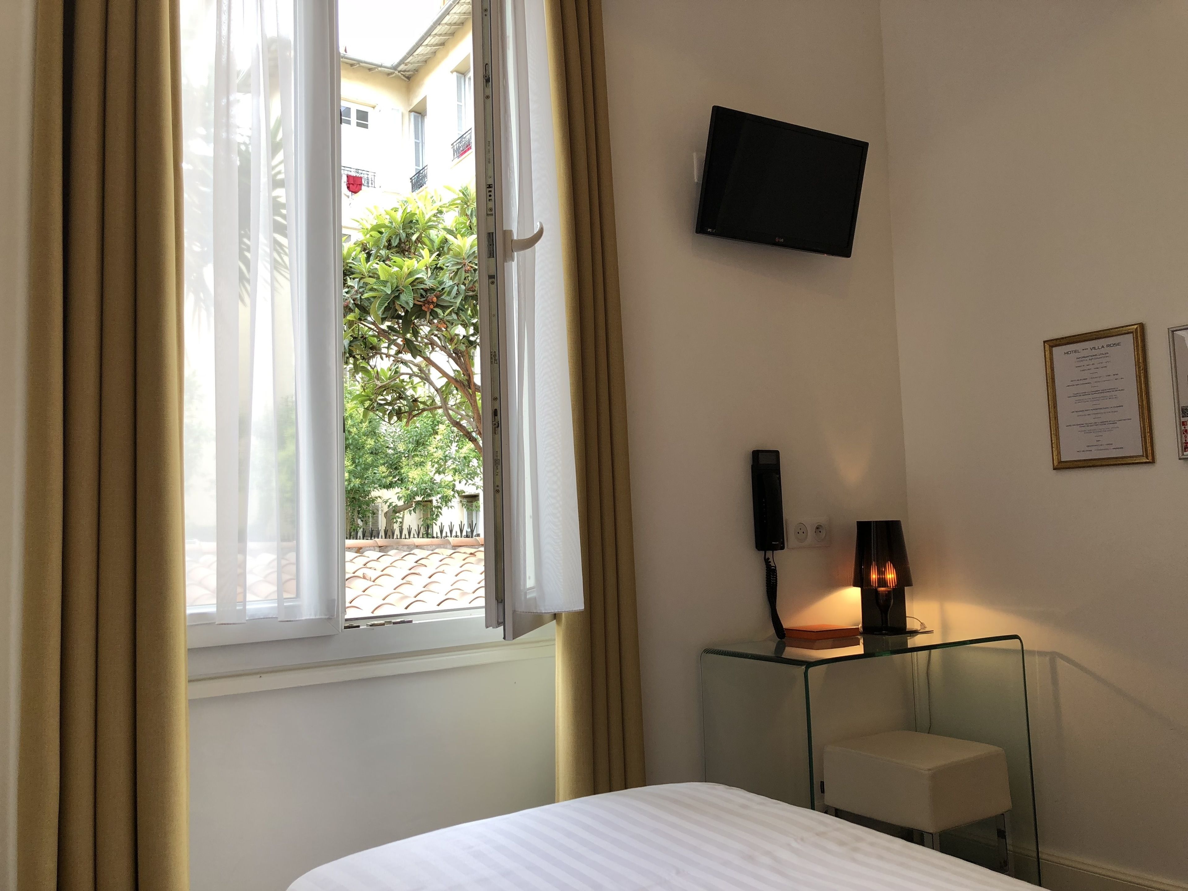 A hotel room with a bed, a TV mounted on the wall, a bedside table with a lamp, and a window with curtains.