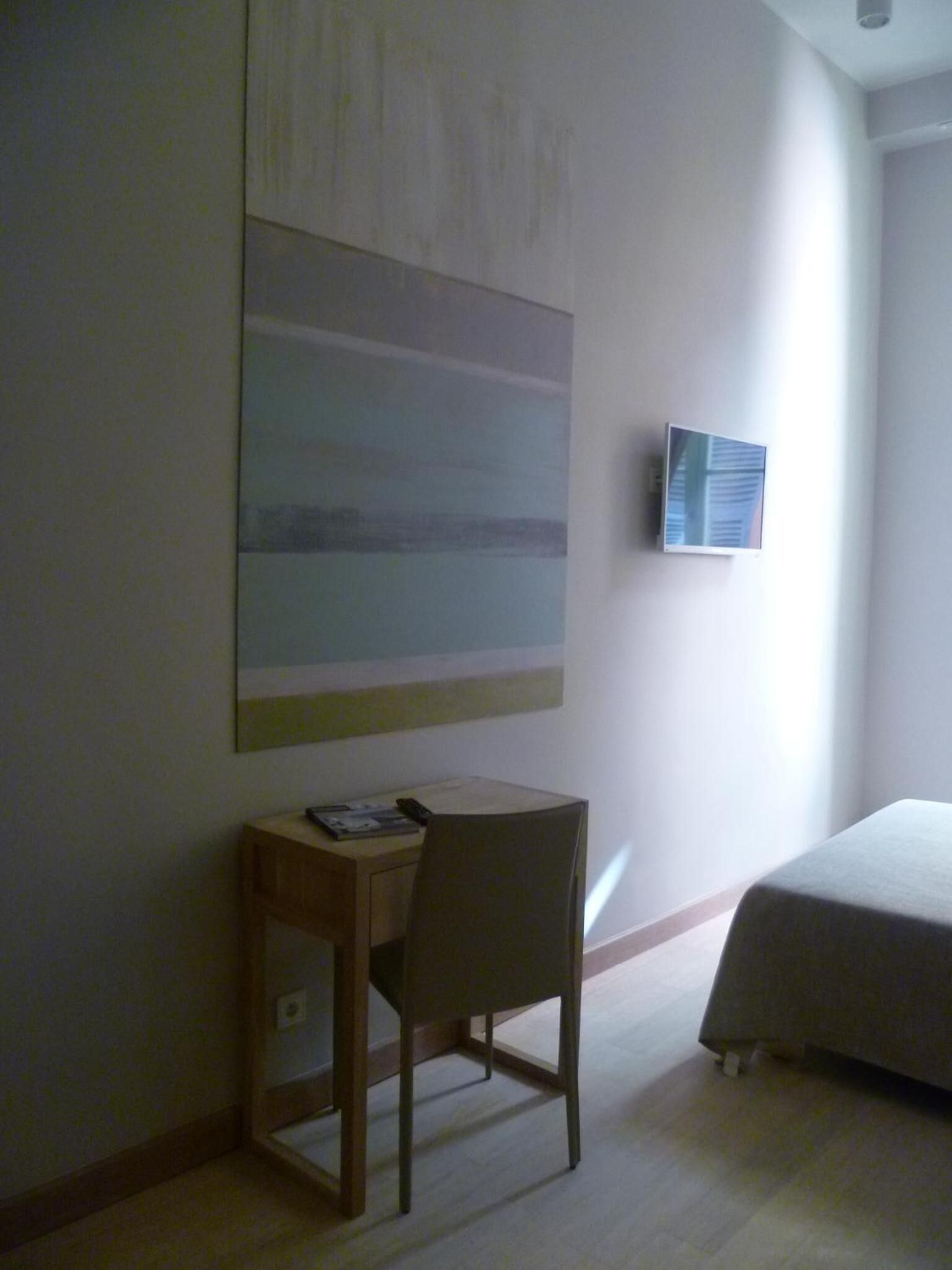 Een hotelkamer met een bed, een bureau met stoel, een groot abstract schilderij aan de muur en een klein raam.